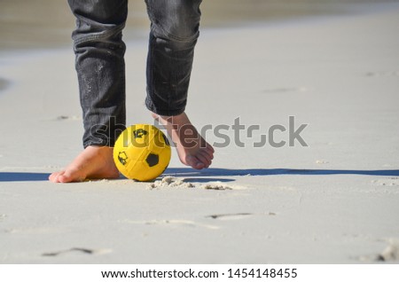 Legs of boy in jeans playing bare foot soccer or football on white sand beach.
