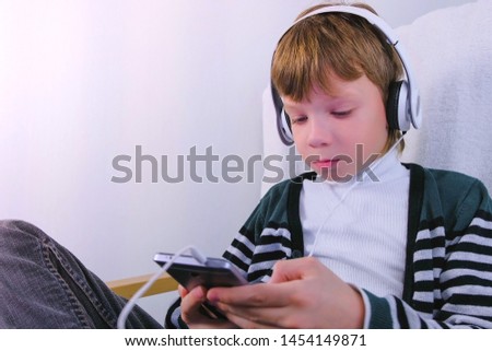 Boy is watching a video in headphones in mobile phone sitting in armchair