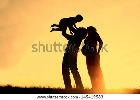 happy family of three people, including a pregnant mother, a father, and a toddler celebrate outside at Sunset, Silhouette against the evening sky