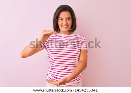 Young beautiful woman wearing striped t-shirt standing over isolated pink background gesturing with hands showing big and large size sign, measure symbol. Smiling looking at the camera. 