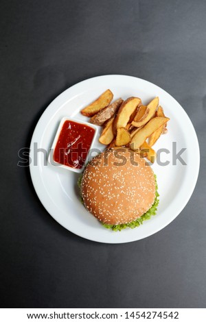 Burger with sesame bun with fried potatoes and ketchup on white plate on black background with copy space. Simple fast food menu with hamburger.