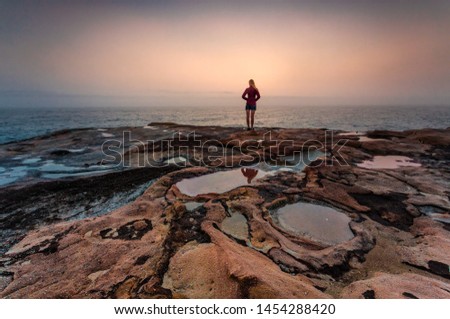 Woman gazing out to sea with foggy coastal sunrise and foreground puddles in weathered sandstone Royalty-Free Stock Photo #1454288420
