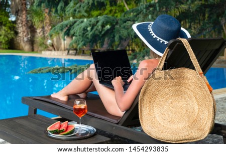 Young woman works on a laptop and drinks cocktail near the pool in a luxury villa