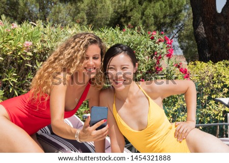 Two beautiful smiling women taking a picture with their cell phones, dressed in bathing suits - Yellow and red bathing suits - Green plants in the background