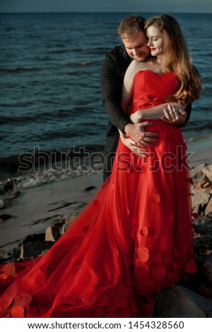 pregnant girl on the beach in a red dress with a long train; pregnant woman on sunset background;  husband with pregnant wife; spouses on the beach
