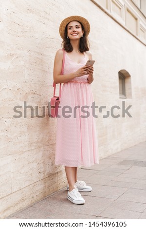 Photo of charming young woman wearing summer dress and straw hat smiling while walking on city street