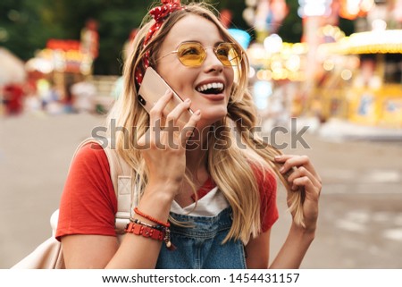 Image of pleased blonde woman wearing girlish clothes talking on cellphone and walking in front of colorful carousel at amusement park