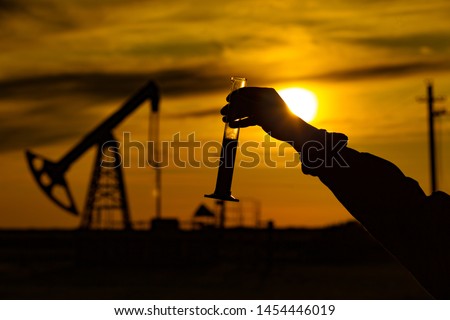 A man holding a flask of oil. Oil rigs in the background. Oil production in Russia. Sunset. Oil pumping Royalty-Free Stock Photo #1454446019