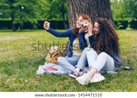 Female students joyful girls taking selfie with adorable puppy in park using smartphone camera having fun together. People, youth and animals concept.