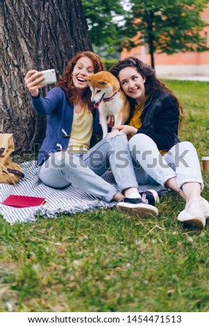 Good-looking curly-haired girls are taking selfie with doggy in park using smartphone camera hugging and stroking animal. People and emotions concept.