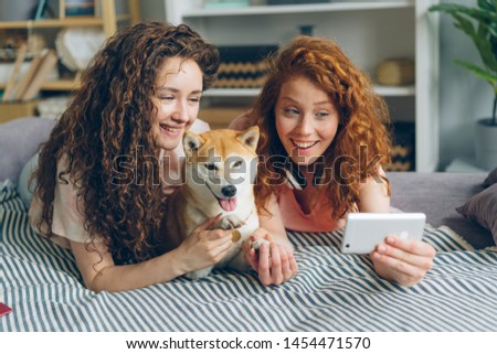 Joyful sisters attractive girls are taking selfie with adorable doggy using smartphone camera lying on couch at home smiling having fun. People and self portrait concept.