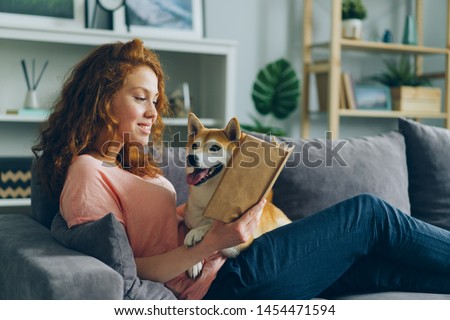 Pretty student young woman is reading book in cozy apartment smiling and petting adorable dog sitting on comfy couch at home. Animals and hobby concept. Royalty-Free Stock Photo #1454471594
