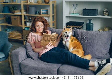 Pretty young woman with long curly red hair is reading book and caressing shiba inu dog sitting on couch at home. Lifestyle, youth culture and animals concept. Royalty-Free Stock Photo #1454471603