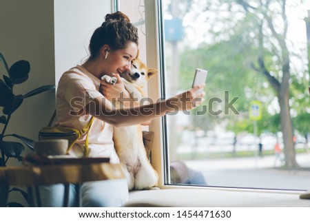 Cheerful lady taking selfie with doggy in cafe using smartphone camera sitting on window sill having fun with cute animal. People and technology concept.