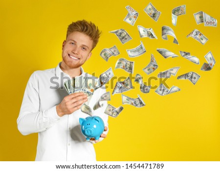 Happy young man with piggy bank and flying money on yellow background