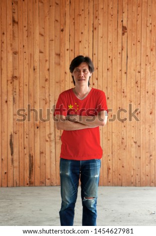Man wearing China flag color shirt and cross one's arm on wooden wall background, large golden star within an arc of four smaller golden stars on red.