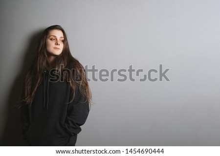 sad young goth woman wearing black sweater leaning against wall with her hands in pockets and looking to side in contemplation - gray background with copy space - adolescence and depression concept.