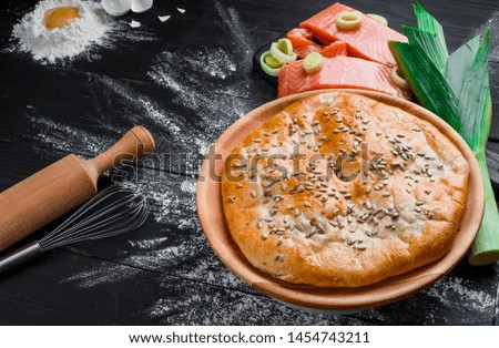 Round homemade salmon pie on round wooden plate on black wooden background in beautiful composition with fresh salmon, broken egg, flour, leek and rolling pin on background. Top view. Space. Close up