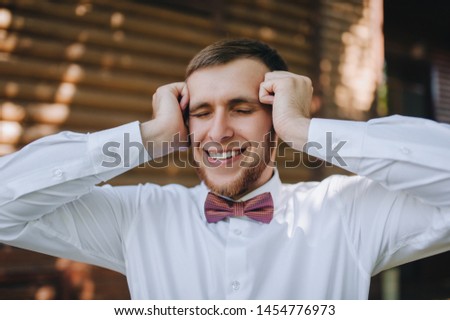 Stylish groom in a white shirt and a red bow-tie holds his head and smiles. Excitement and experience. Wedding portrait close-up of a young, handsome man. Photography and concept.