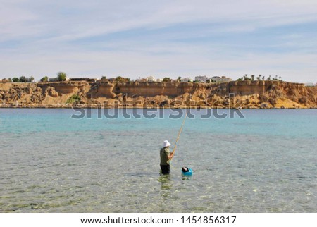 Fisherman with a fishing rod in the azure sea, in the distance yellow rock Royalty-Free Stock Photo #1454856317