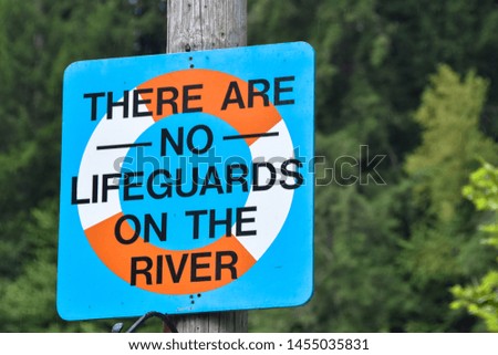 blue sign with life preserver red and white reading there are no lifeguards on the river signage on a wood pole green tree background 