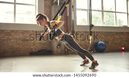 Working hard. Side view of young athletic woman with perfect body in sportswear doing push ups with trx fitness straps in the gym Royalty-Free Stock Photo #1455092690