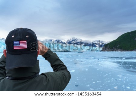 Taking a picture of Hubbard Glacier, Man is wearing a cap with use flag. Expedition to the glaciers of Alaska. Green mountain and snowy glacier looks amazing. It is summer time cruise.