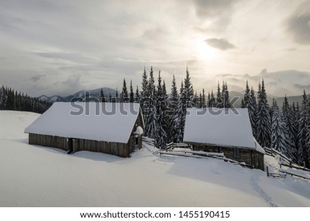 Winter mountain landscape. Old wooden houses on snowy clearing on background of mountain ridge, spruce forest and cloudy sky. Happy New Year and Merry Christmas card.