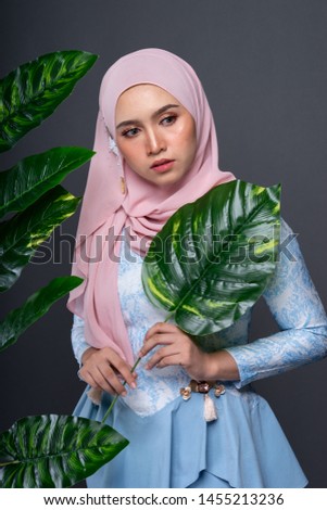 Beautiful female model wearing peplum dress with hijab, a modern lifestyle outfit for Muslim woman, posing with artificial leaves isolated over grey background. Studio hijab fashion and beauty concept