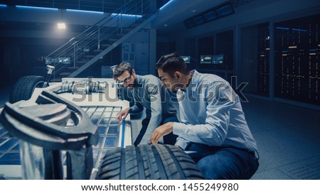 Two Automotive Engineers Working on Electric Car Chassis Platform, Taking Measures, working with 3D CAD Software, Analysing Efficiency. Vehicle Frame with Wheels, Engine and Battery. Royalty-Free Stock Photo #1455249980
