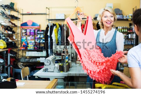Portrait of professional positive mature female tailor showing dress to customer at counter in sewing atelier