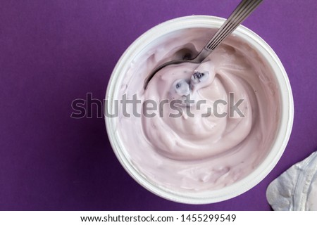 Yogurt cup with blue berry yoghurt, spoon and foil lid isolated on purple background with text space - top view photograph of creamy fruit yoghurt Royalty-Free Stock Photo #1455299549