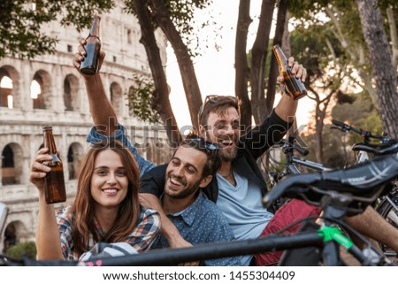 Three happy young friends tourists on a bench at Colosseum in Rome having fun drinking and toasting with beers at sunset