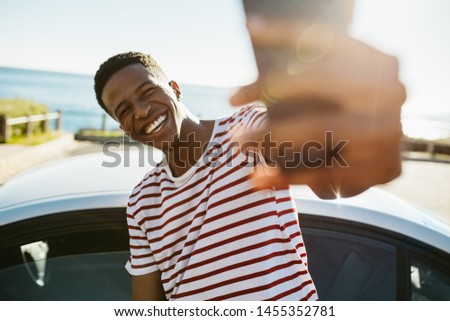 Smiling young african man taking selfie while standing by a car outdoors. African american guy making a self portrait outdoors on a summer day.