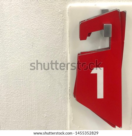Number one, on the wall is a white sign. On a white background, the number "1" is colored red. Concept - number one, best, first. Plate with the number next to the fitting room in a clothing store.