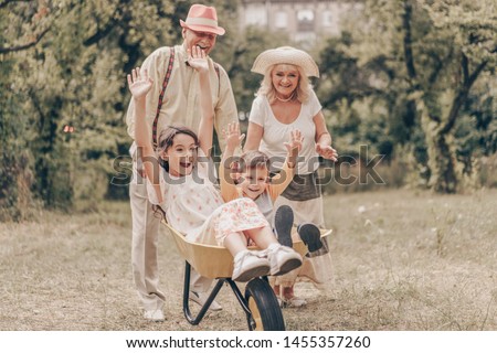 Grandparents in park with grandchildren playing with cart. Family have fun in garden. Old people happy to see granddaughter and grandson Royalty-Free Stock Photo #1455357260