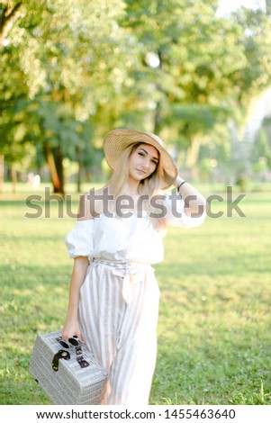 Young beautiful girl walking in garden and keeping bag, sunglasses and hat. Concept of walking in park and summer fashion.