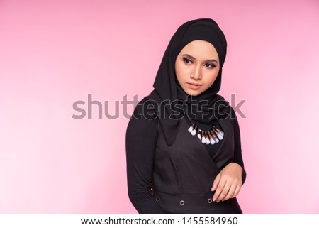 Portrait of a beautiful Muslim female model wearing black shirt with hijab isolated over pink background. Studio fashion and beauty make up concept.