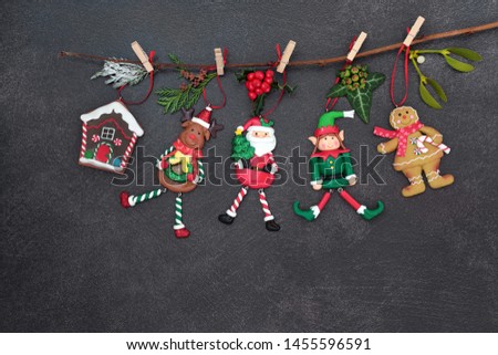 Christmas tree decorations hanging on a branch with winter flora and baubles of santa claus, reindeer, elf, gingerbread house and cookie lady on dark grey grunge background with copy space.