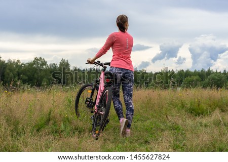 Young woman cycling pink bike. Happy fitness woman stands with a bicycle on green field at cloudy day. Slim girl wearing pink clothes rides a bicycle outdoors. Sport  Activity Healthy lifestyle