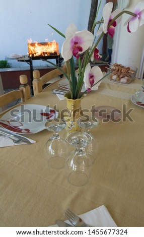 Decorated table in the garden. Dishware Decor Dinner Concept. Dishes ready and clean water cups. Barbeque in summer time. Flower pot center. Fire in bbq