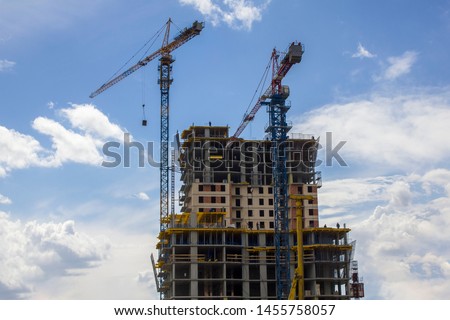 two tower cranes on top of a skyscraper under construction with workers against a blue sky with clouds