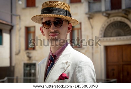 Young dandy wearing a tie, a red pocket handkerchief and a straw boater hat, taking a stroll in the streets of an Italian town Royalty-Free Stock Photo #1455803090
