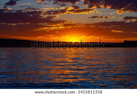 Last light of day over Wahweap Bay in Lake Powell, Arizona.  The sun is sinking behind the distant mountain range and the sky goes from yellow and orange to blue through the clouds and reflects below.