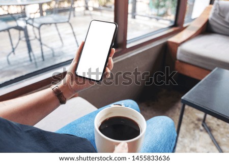 Mockup image of woman holding black mobile phone with blank desktop screen while drinking coffee in cafe