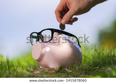 Pink piggy bank with glasses on grass and hand putting in a coin