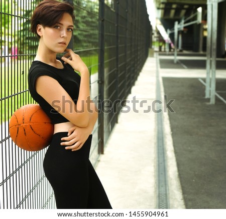 Sporty caucasian girl playing basketball - short haircut girl in a sporty wear. Portrait of a young woman holding a basketball ball, Girl at Basketball court behind fence mesh netting, outdoors