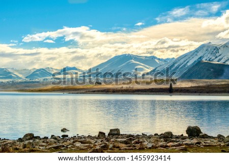 Snowy mountains and pine trees, cars running on gravel road causing orange dust on the lake of Tekapo New Zealand in the morning. The sky is blue with beautiful clouds.