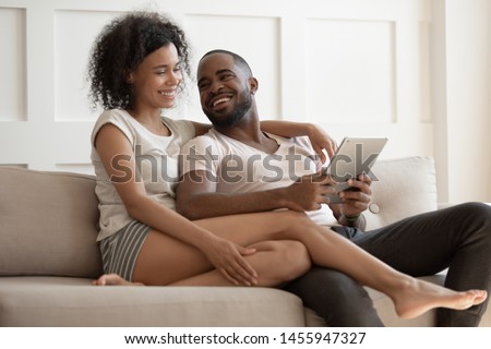 Happy young black couple enjoying using wireless digital tablet sit on sofa on leisure, smiling african american husband and wife holding pc computer doing shopping online or reading e-book at home