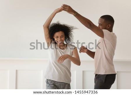 Happy funky young african american romantic couple holding hands dancing laughing at home, carefree active millennial black husband and wife wearing pajamas having fun bonding enjoy dance in bedroom